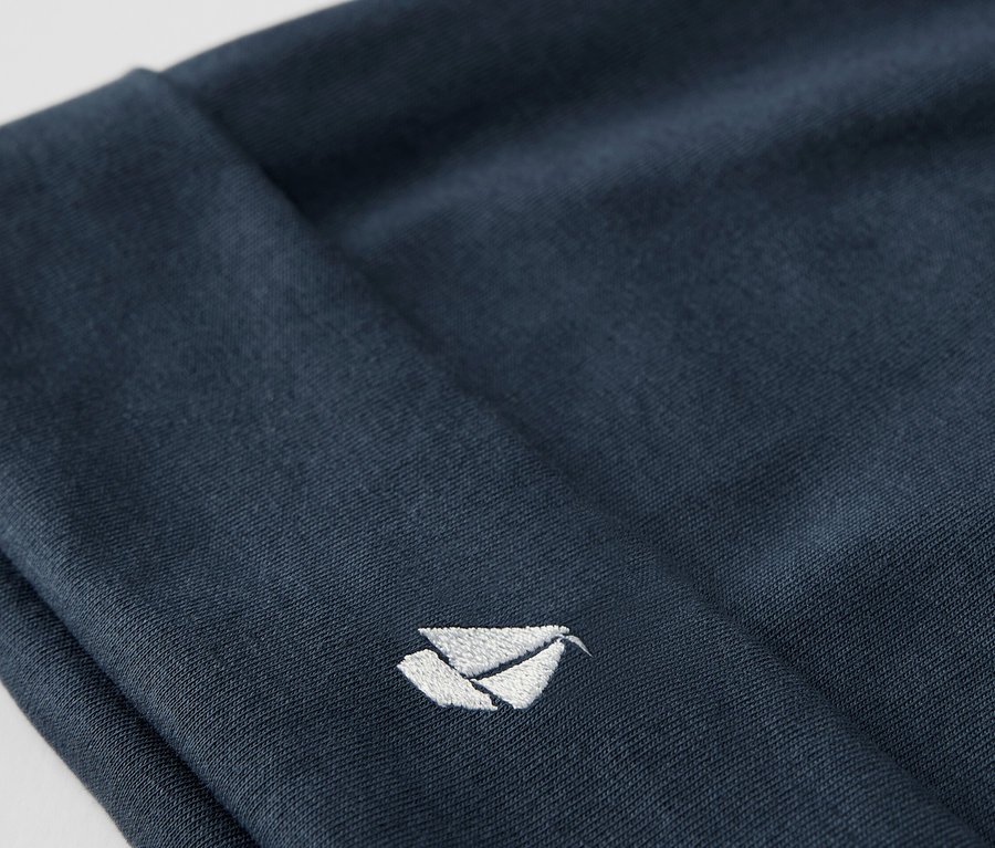 Detailaufnahme einer blauen Stoffmütze aus einem Baby-Set, mit einem aufgestickten weißen Logo.