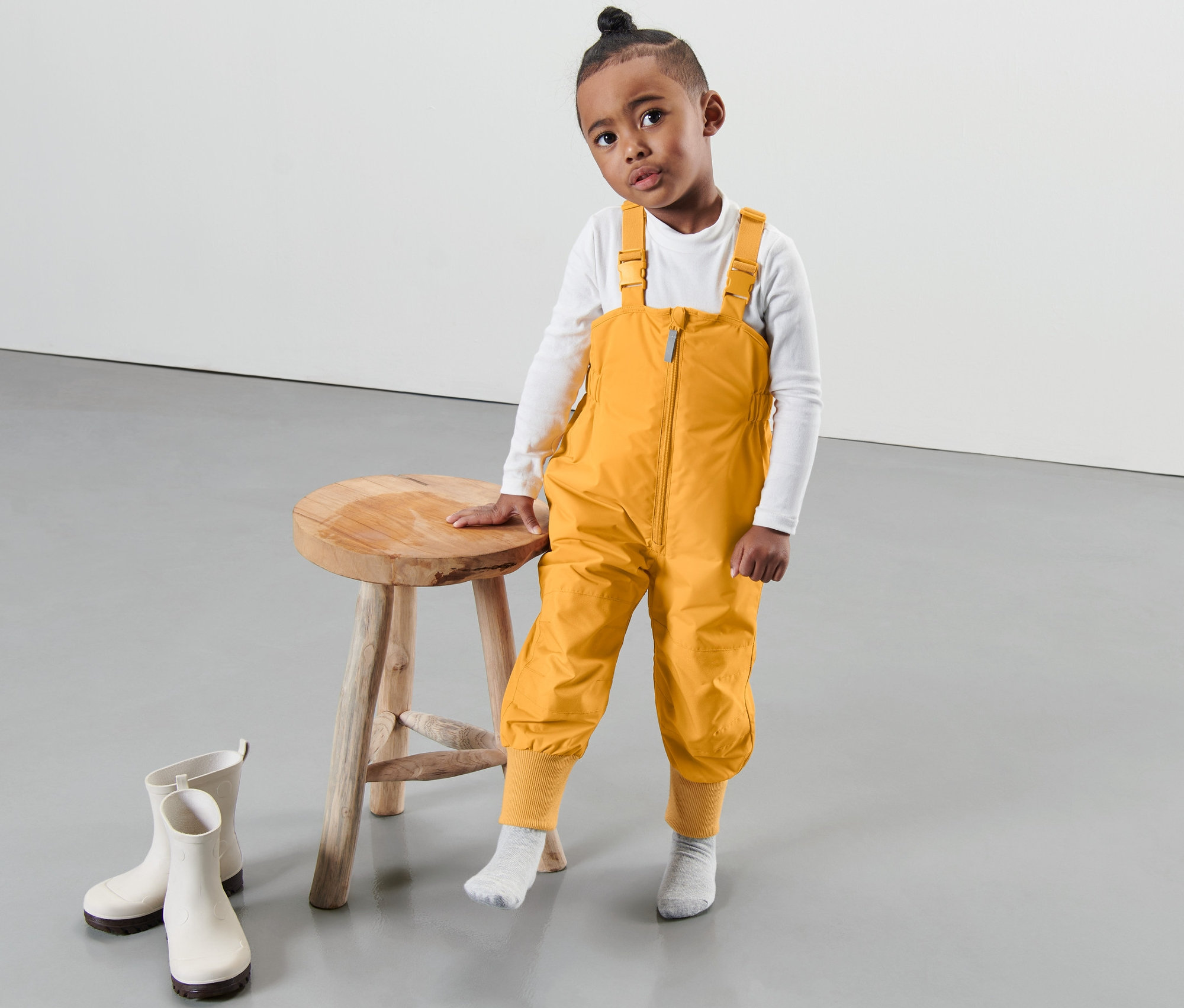 Ein Junge in einer korngelben Kleinkind-Thermo-Regenhose steht neben einem Hocker, Gummistiefel liegen auf dem Boden.