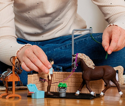 Eine Person mit roten Haaren spielt auf einem Holzboden mit dem Schleich »Horse Club« Waschplatz. Sie hält eine kleine Mädchenfigur, die ein braunes Pferd an einem lila Halfter am Waschplatz pflegt. Daneben steht ein Sattelständer mit Sattel.