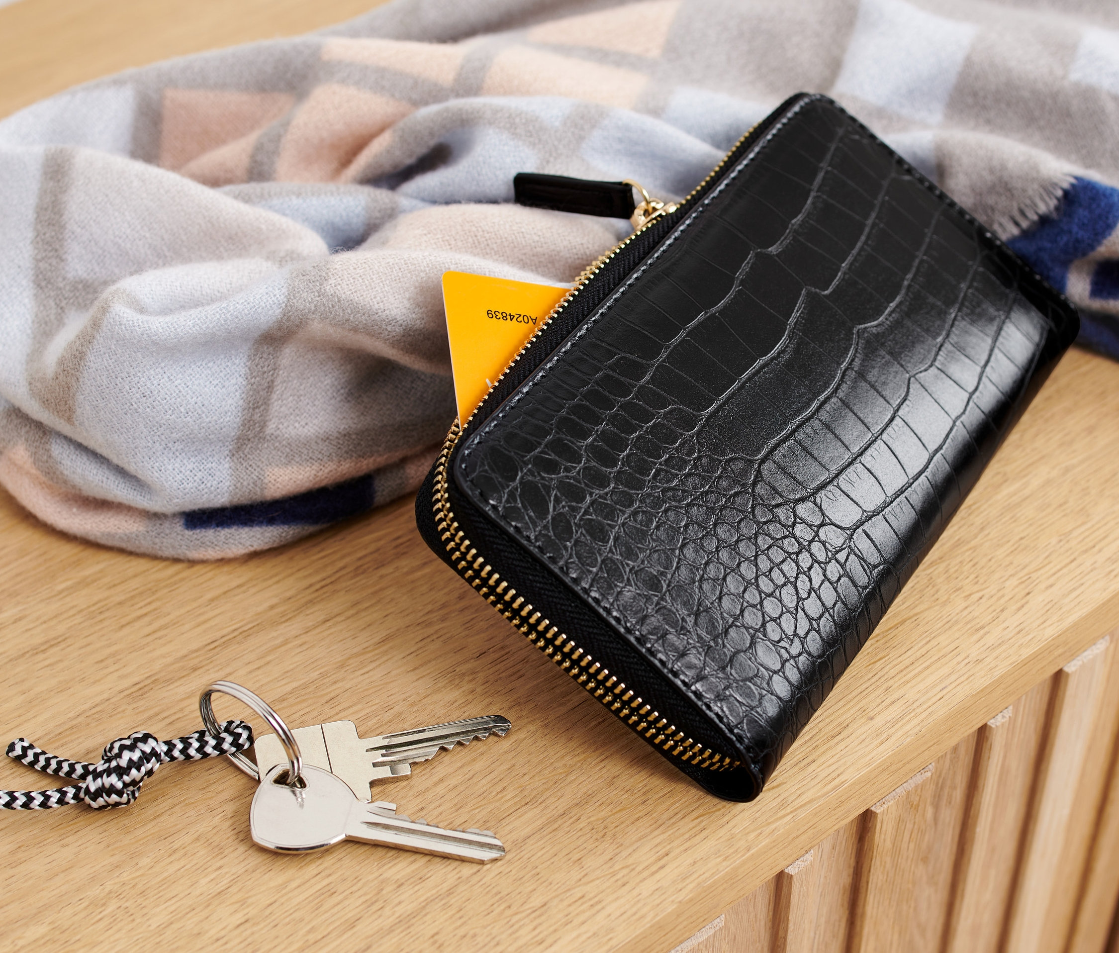 Eine schwarze Geldbörse liegt auf einer Holzoberfläche neben Schlüsseln und einem gemusterten Schal.