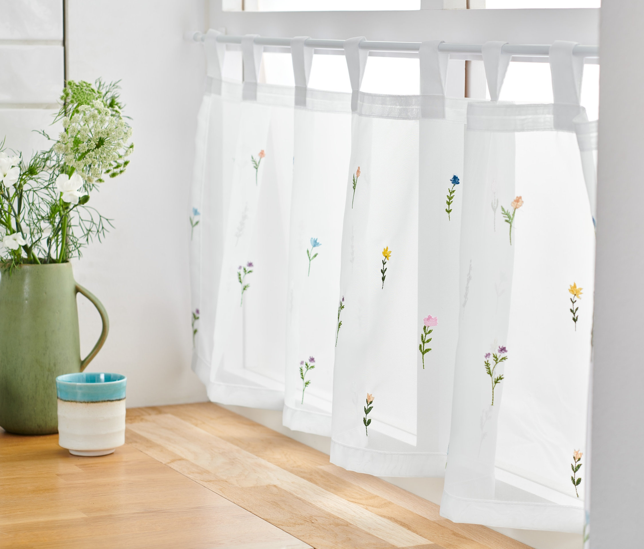 Eine bestickte Bistrogardine mit Blumenmuster hängt an einem Fenster.