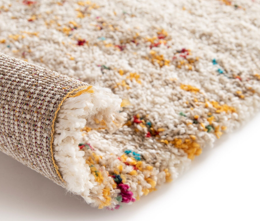 Detailansicht eines beigen Teppichs »Himalaya« mit Farbtupfern, teilweise aufgerollt, der die Unterseite zeigt.