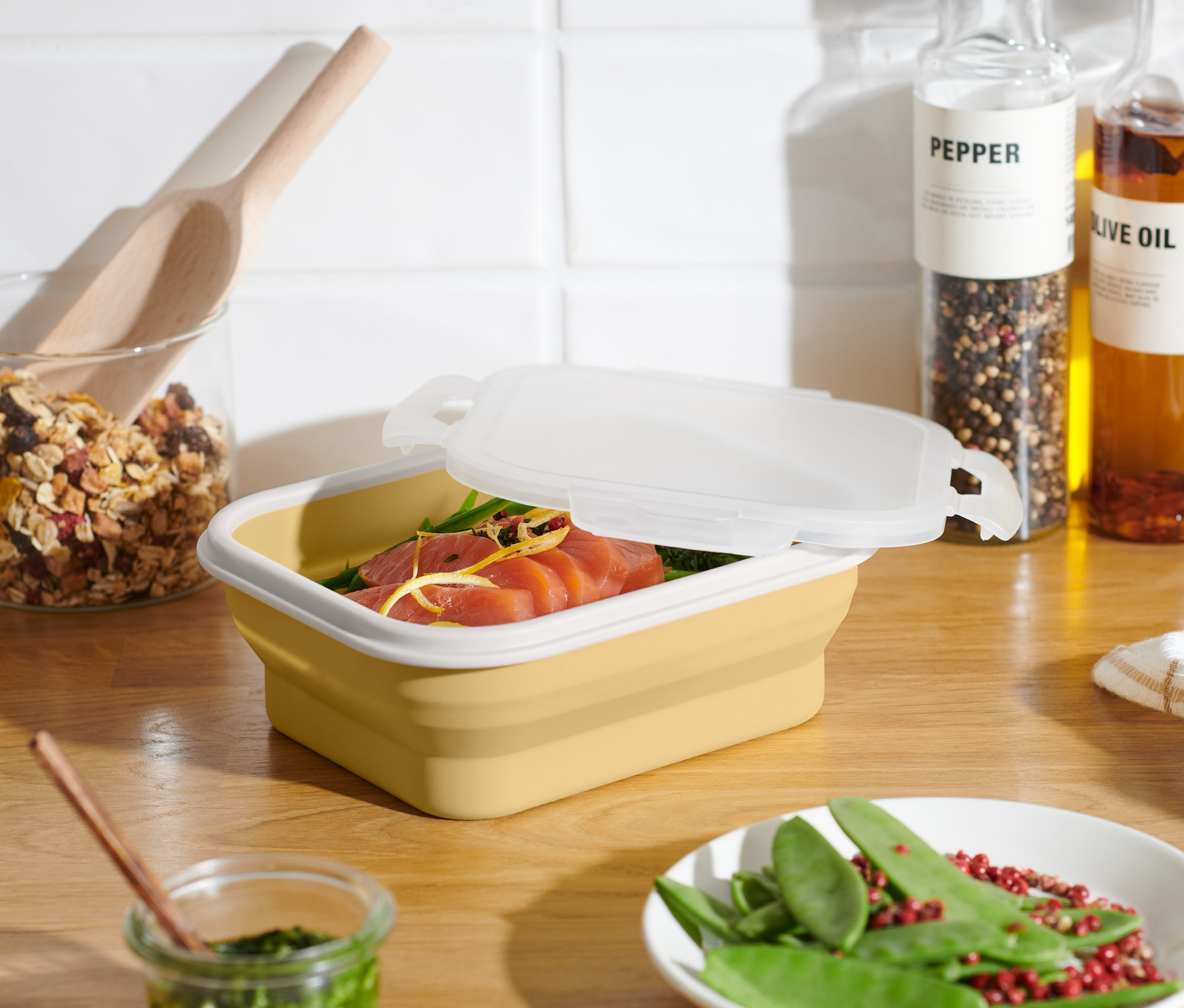 Gelbe, faltbare Frischhaltedose mit leicht geöffnetem Deckel, gefüllt mit Lachs. Auf dem Tisch liegt ein Teller mit grünen Erbsen und rotem Pfeffer.