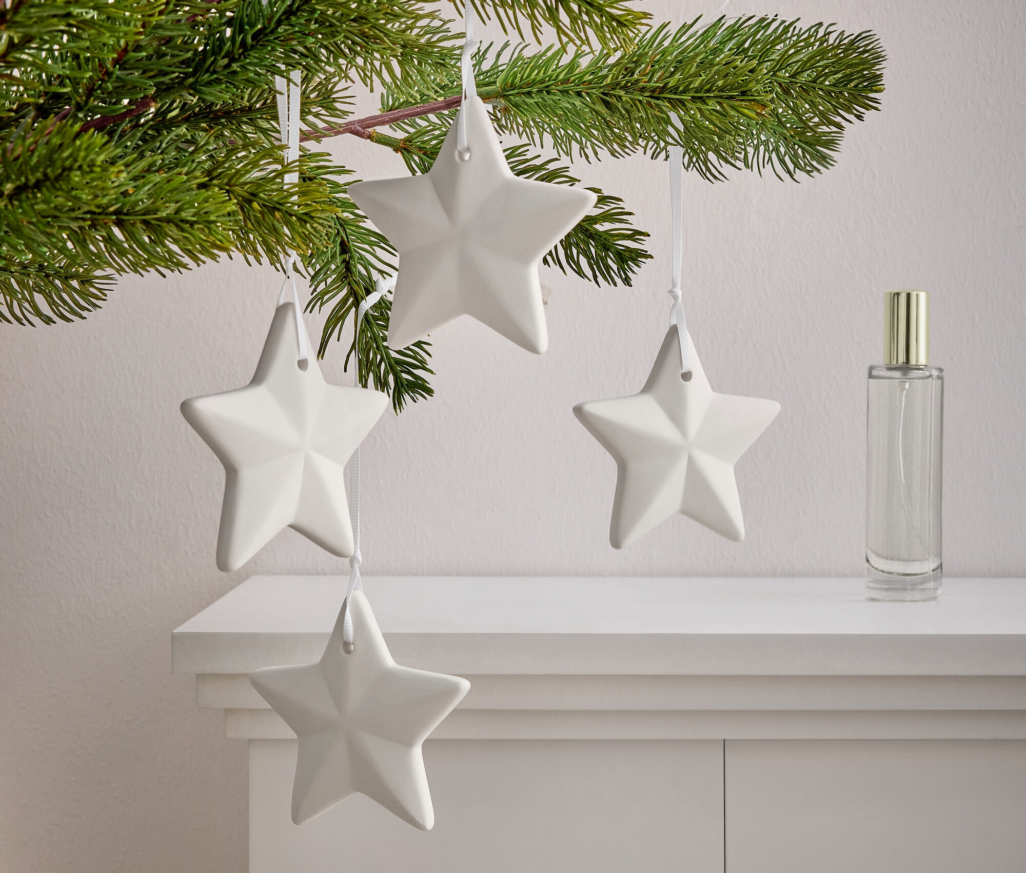 Ein Zweig mit Nadeln, an dem vier weiße Sterne an einem weißen Schrank hängen. Neben den Sternen steht eine Parfümflasche.