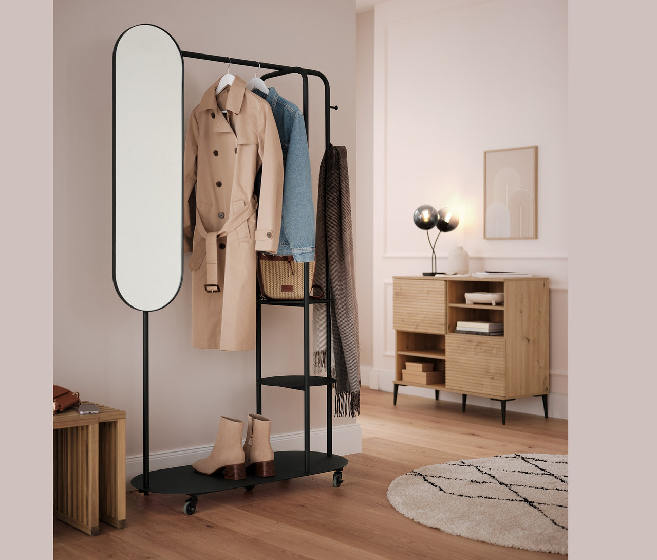 Schwarzer Kleiderständer mit ovalem Spiegel und Ausziehhocker. An der Stange hängen ein beiger Mantel, eine blaue Jacke und ein grauer Schal.
