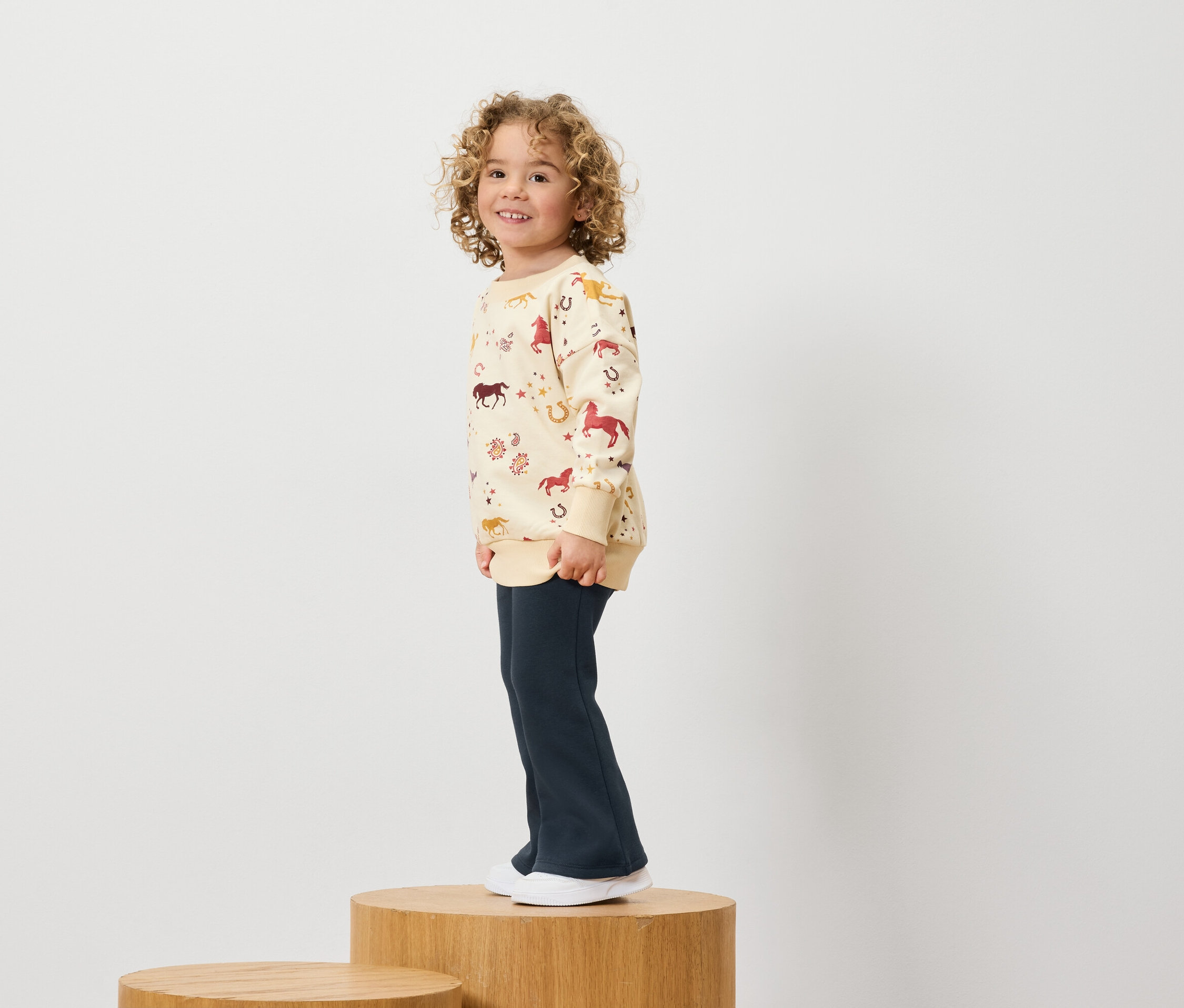 Junge mit lockigem Haar, trägt ein Sweatshirt mit Pferdeaufdruck und dunkelblaue Hosen, steht auf einem Holzsockel.