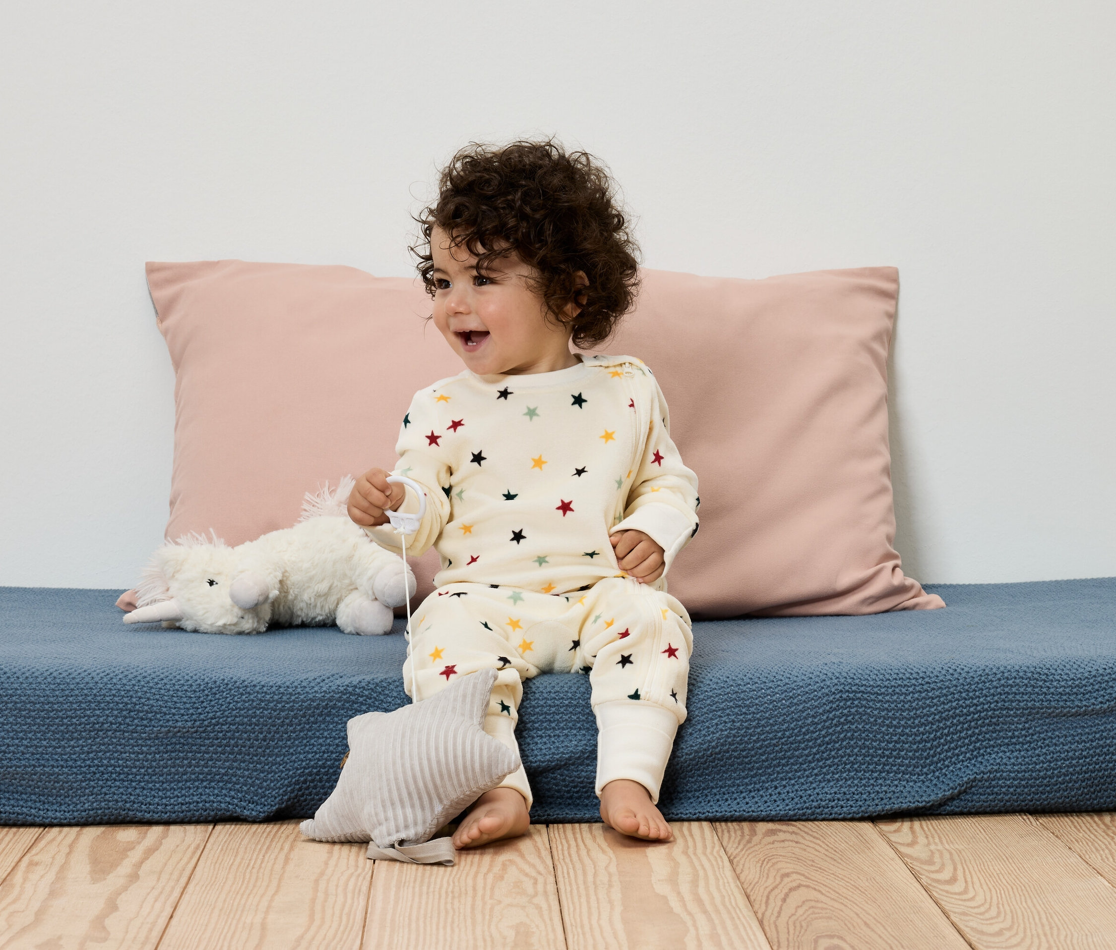 Ein Kind sitzt auf einer blauen Unterlage mit Sternenpyjama, rosa Kissen und Spielzeug.
