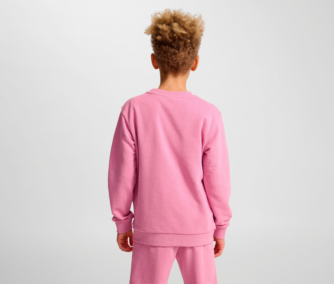 Ein Junge mit lockigem Haar steht mit dem Rücken zur Kamera und trägt ein rosa HUMMEL HMLJR Base-Sweatshirt.