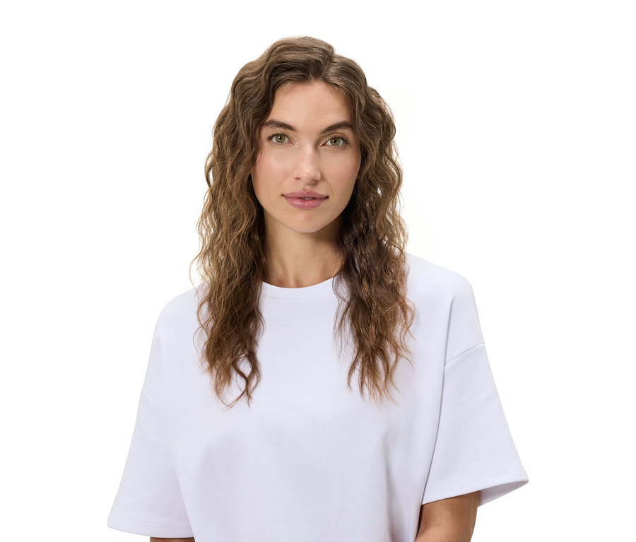 Porträt einer Frau mit gewelltem braunem Haar und einem weißen T-Shirt vor einem weißen Hintergrund.