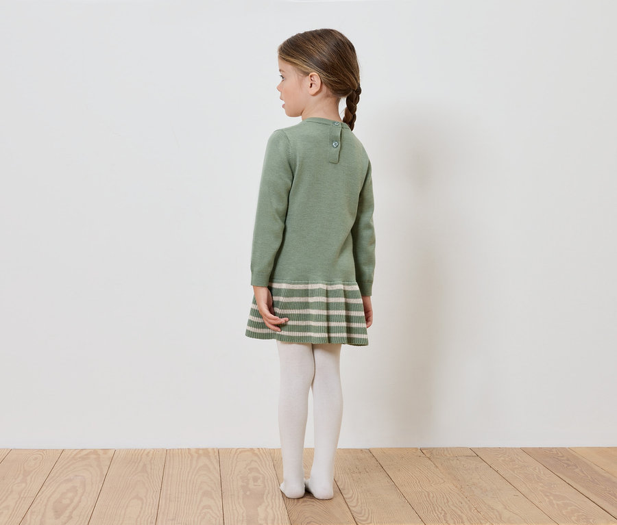 Mädchen in grünem Strickkleid steht mit dem Rücken zur Kamera.