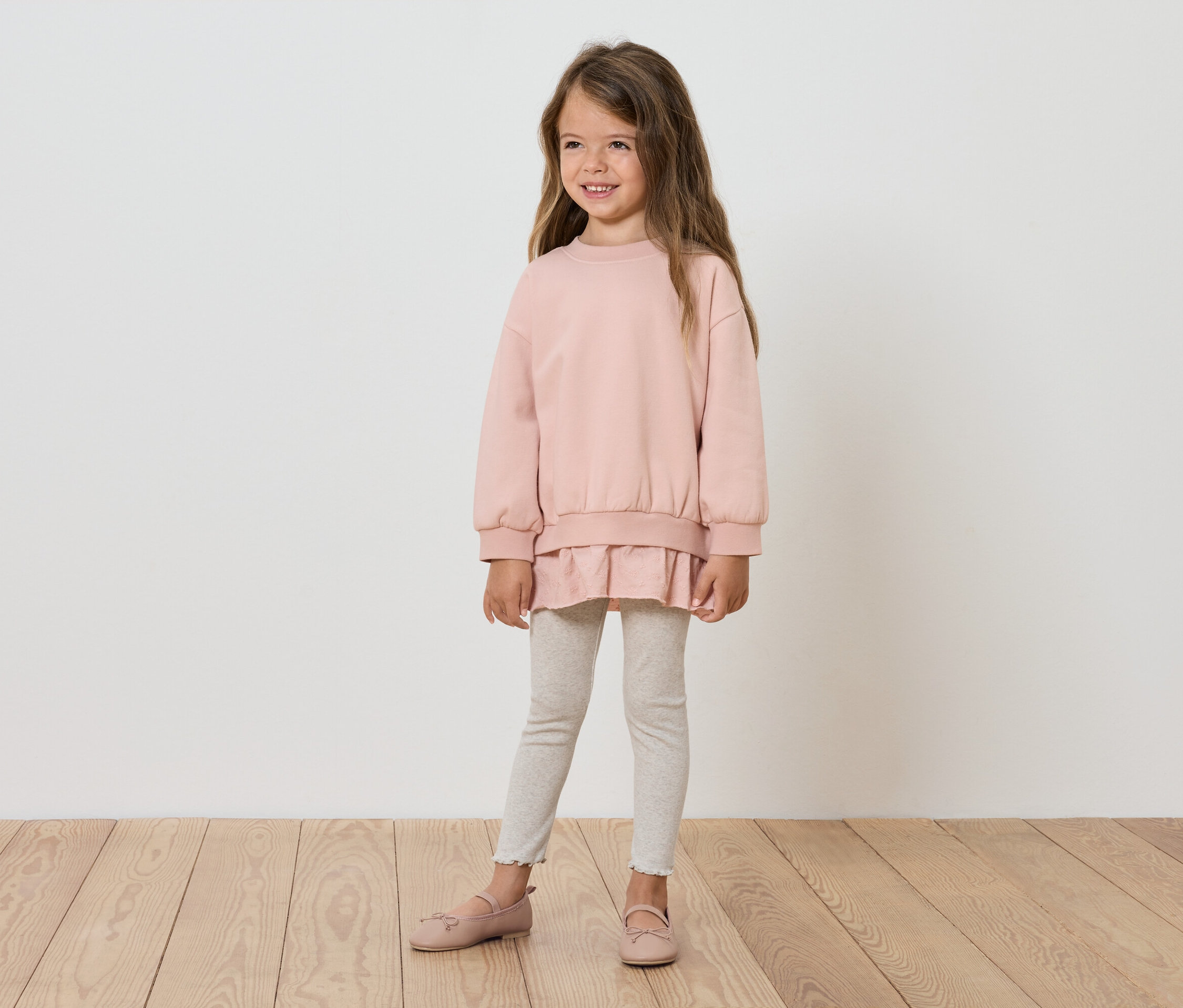Mädchen steht und lächelt, trägt einen rosa Pullover, weiße Leggings und rosa Schuhe.