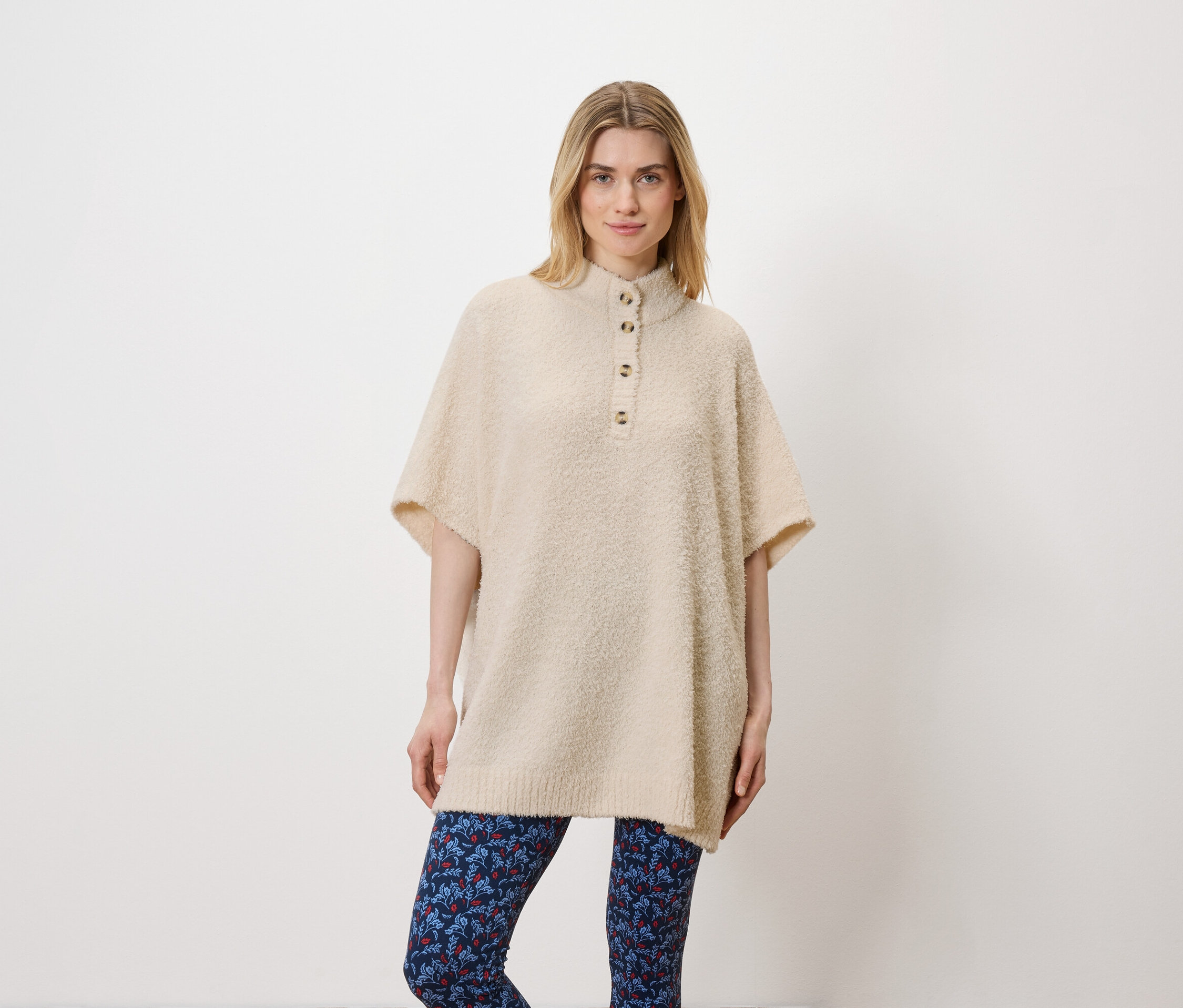 Eine Frau posiert im Studio vor einem weißen Hintergrund. Sie trägt einen beigen Pullover und blaue Leggings.