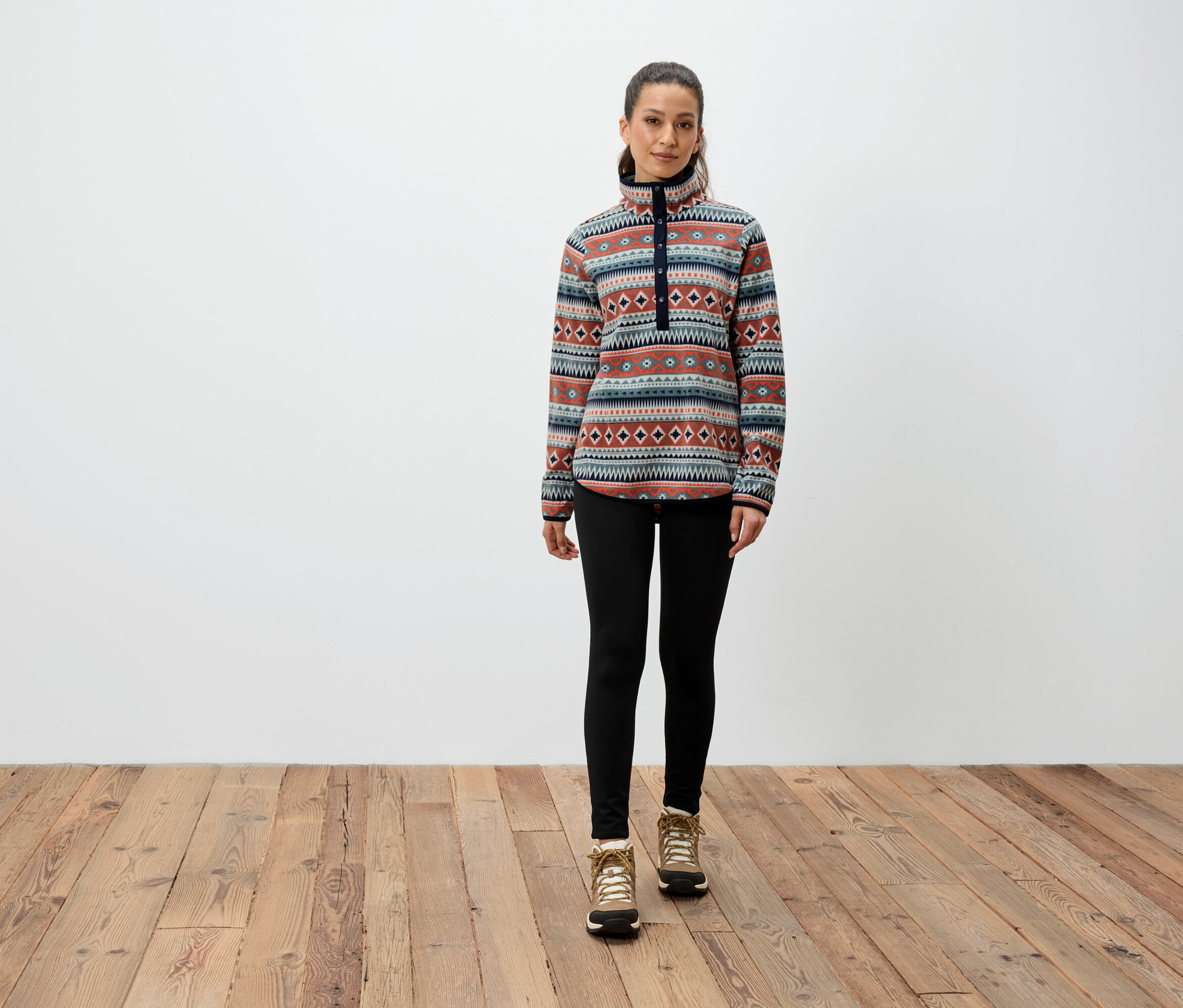 Eine Frau steht auf einem Holzboden und trägt ein gemustertes Sweatshirt, schwarze Leggings und braune Stiefel.