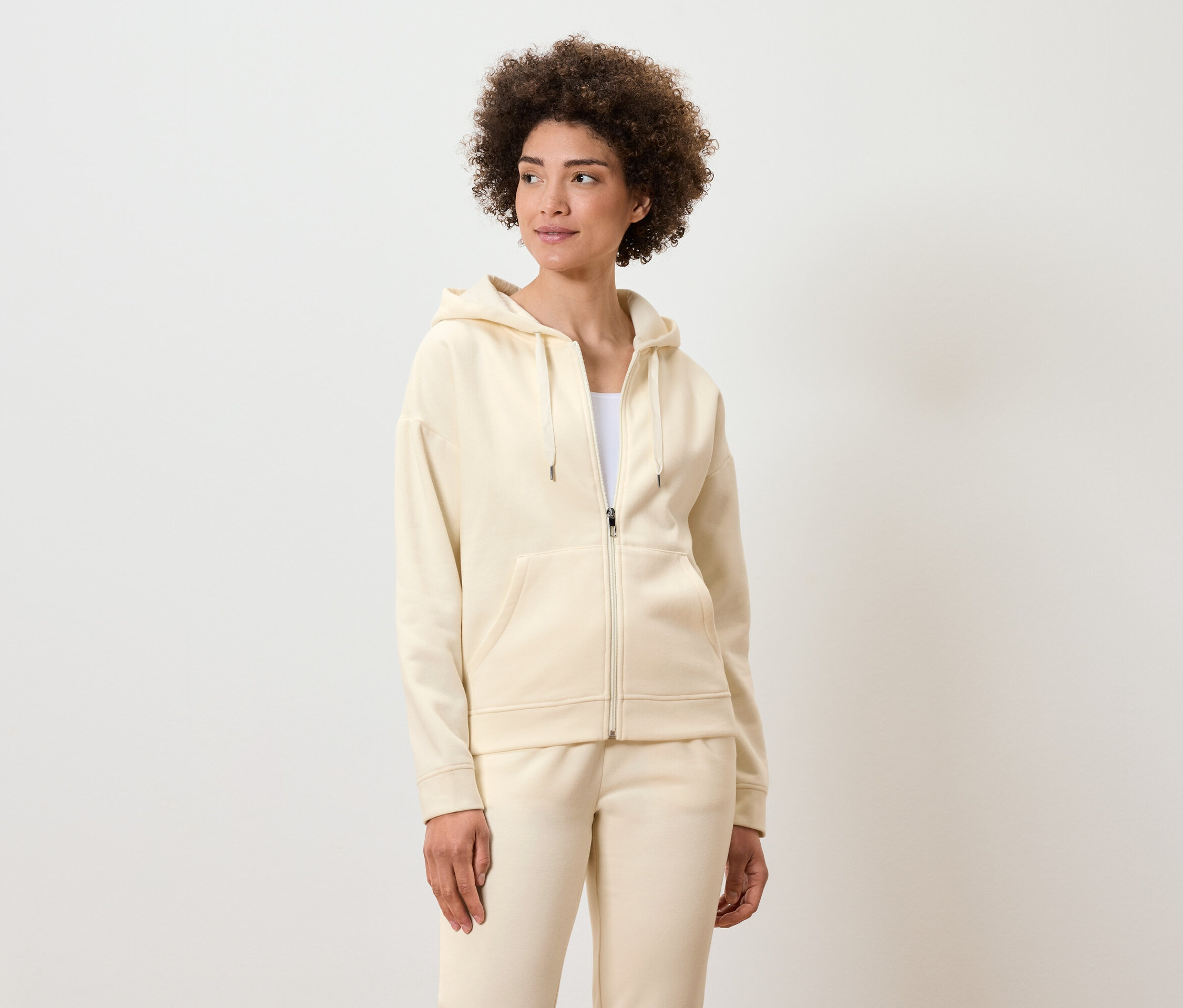 Frau mit lockigem Haar posiert in einem cremefarbenen Reißverschluss-Hoodie und Trainingshose.
