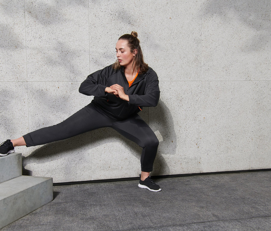 Frau in Sporthose und Sportjacke dehnt sich an einer Wand. Eine Wasserflasche steht neben ihr.