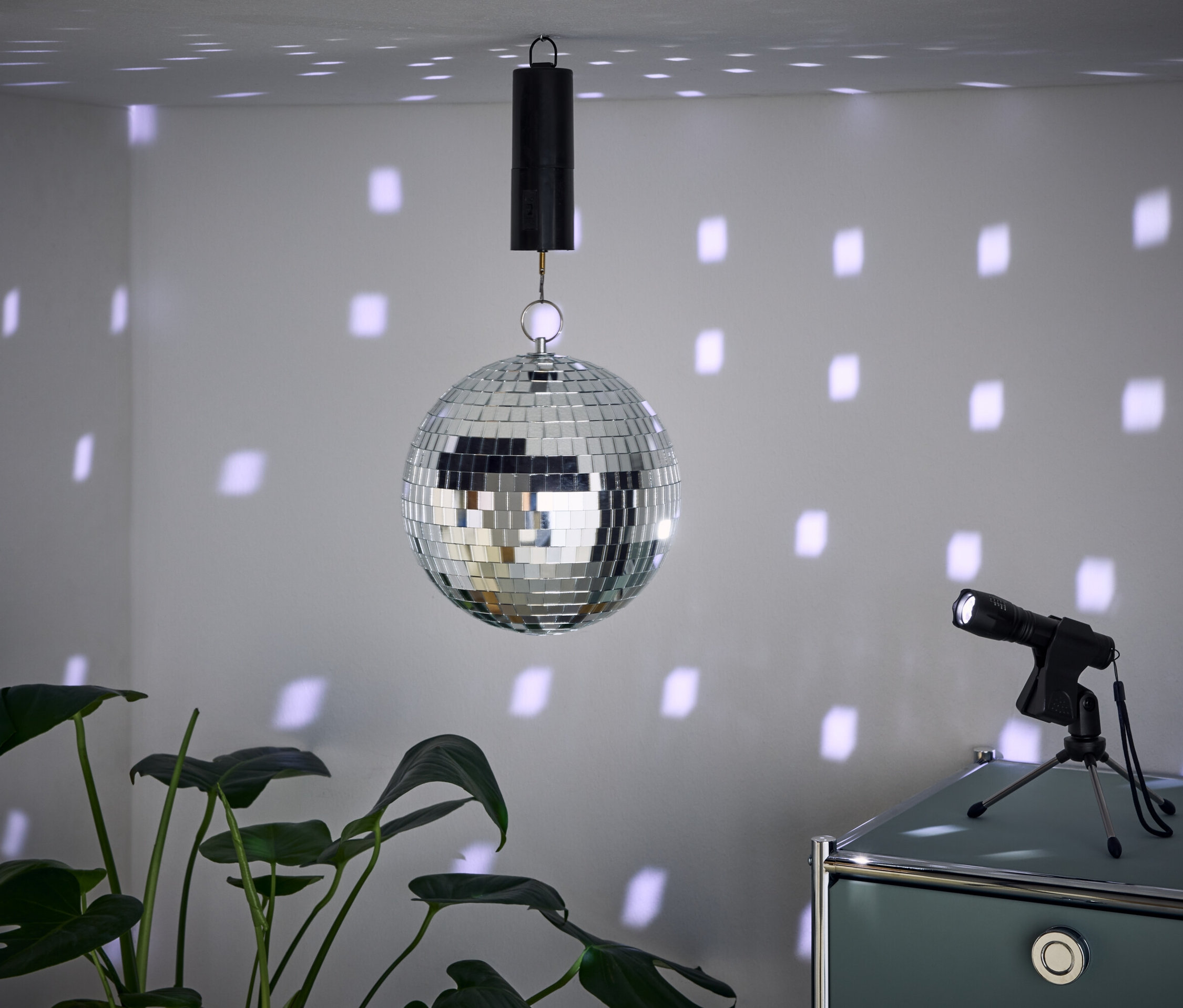 Eine Spiegelkugel hängt von der Decke und wirft Lichtflecken an die Wände. Daneben steht ein Scheinwerfer auf einem Schrank und im Vordergrund ist eine Pflanze.