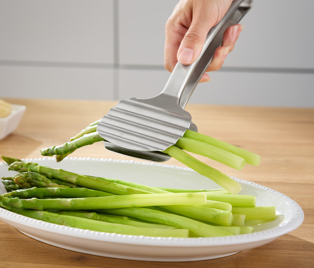 Eine Hand hält eine silberne Spargelzange über eine Schale mit grünem Spargel.