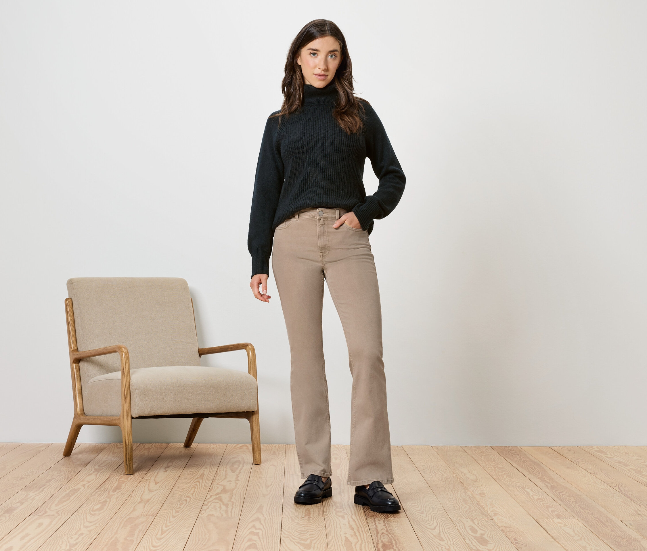 Frau im schwarzen Rollkragenpullover und beigen Hosen posiert neben einem beigen Sessel.