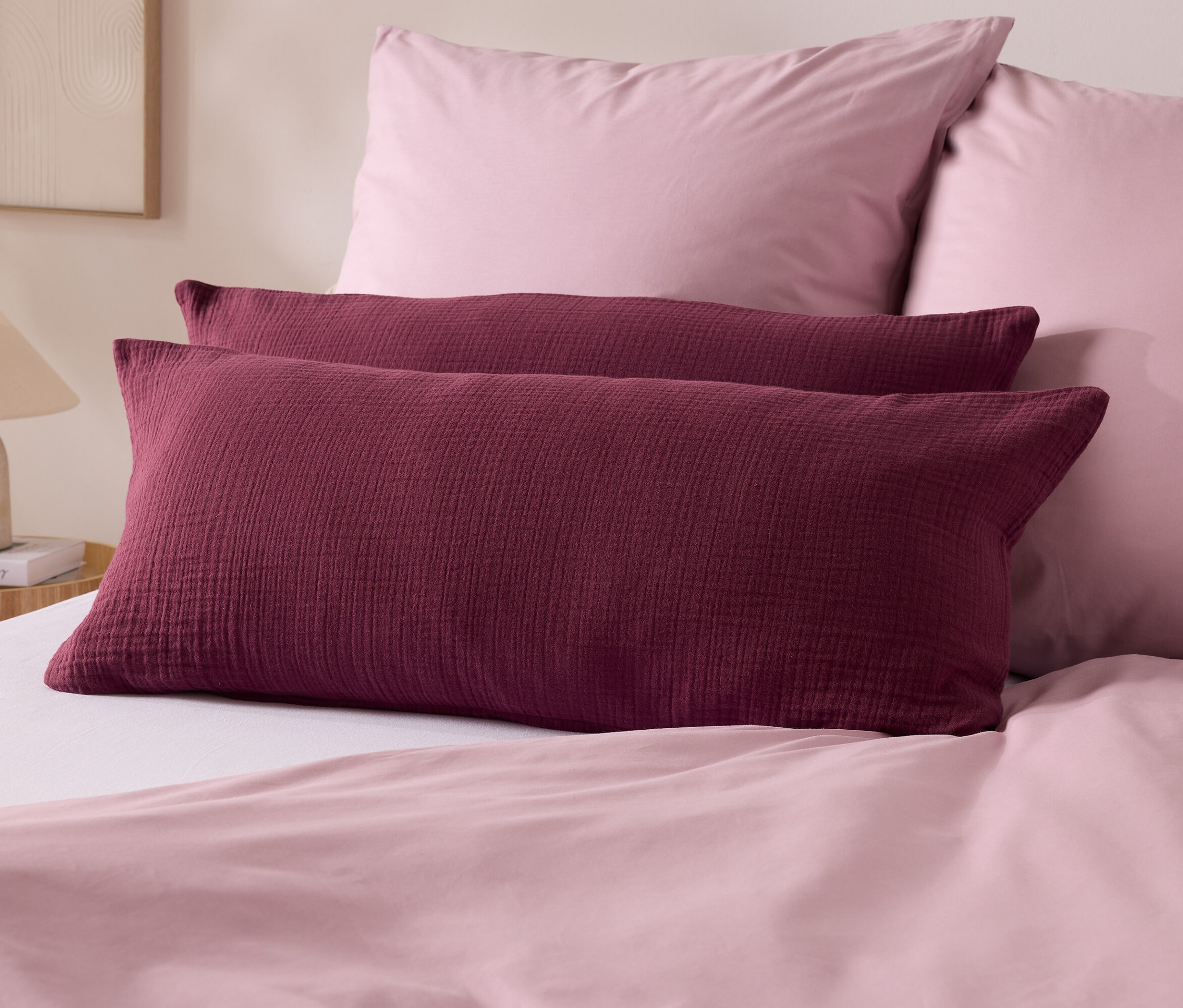 Zwei bordeauxrote Kissen liegen auf einem Bett mit rosa Bettwäsche. Im Hintergrund befinden sich zwei rosa Kissen und eine Lampe.