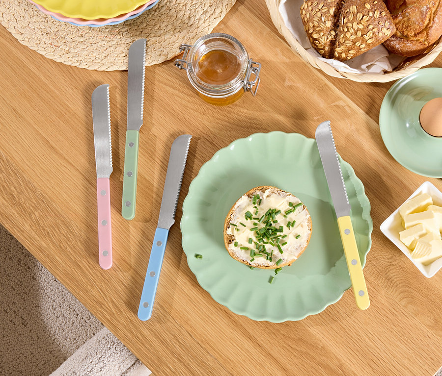 Auf einem Holztisch liegen ein Teller mit Brot mit Butter und Kräutern, Butter in einer Schüssel, ein Ei in einem Eierbecher und Messer mit bunten Griffen.