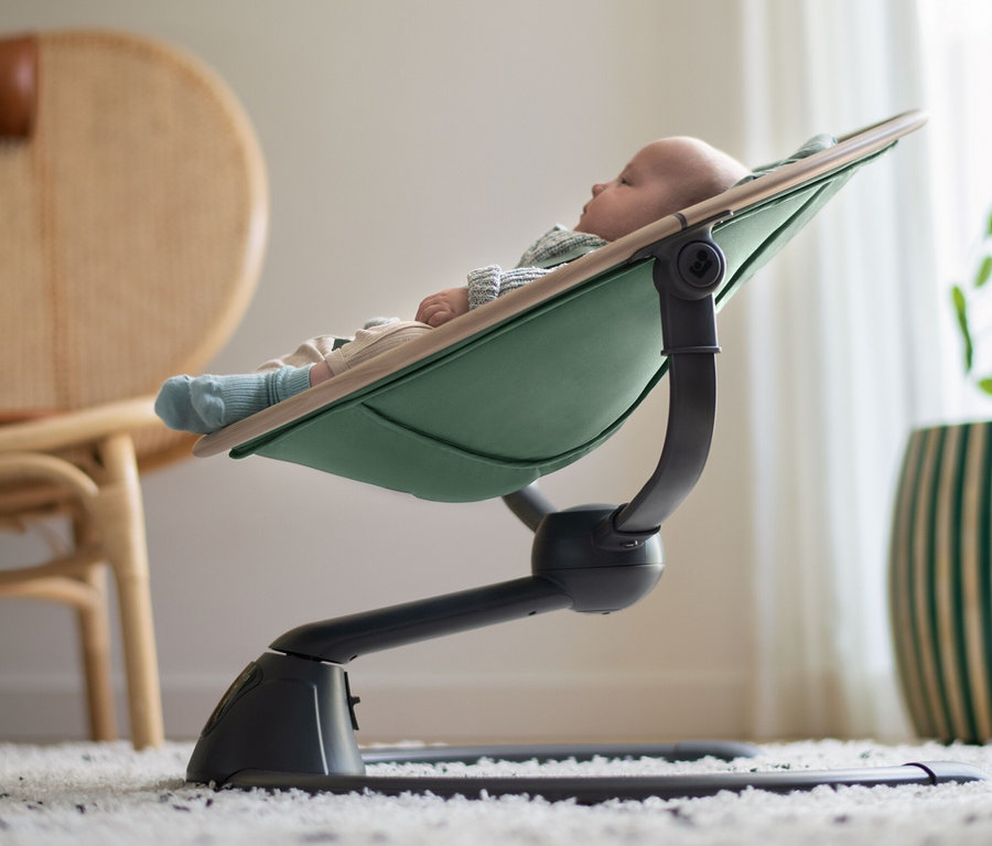 Ein Baby liegt in der Maxi-Cosi Elektrische Schaukel »Cassia«, im Hintergrund ein Korbsessel und ein Blumentopf.