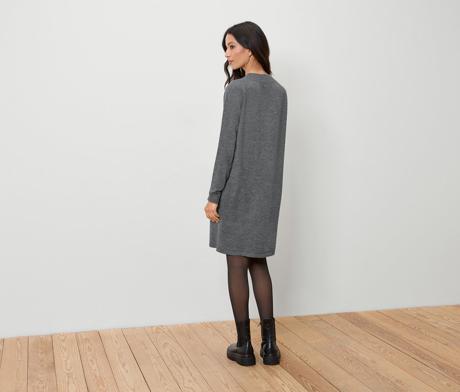 Eine Frau steht mit dem Rücken zur Kamera und trägt ein graues Kleid, eine schwarze Strumpfhose und schwarze Stiefel.