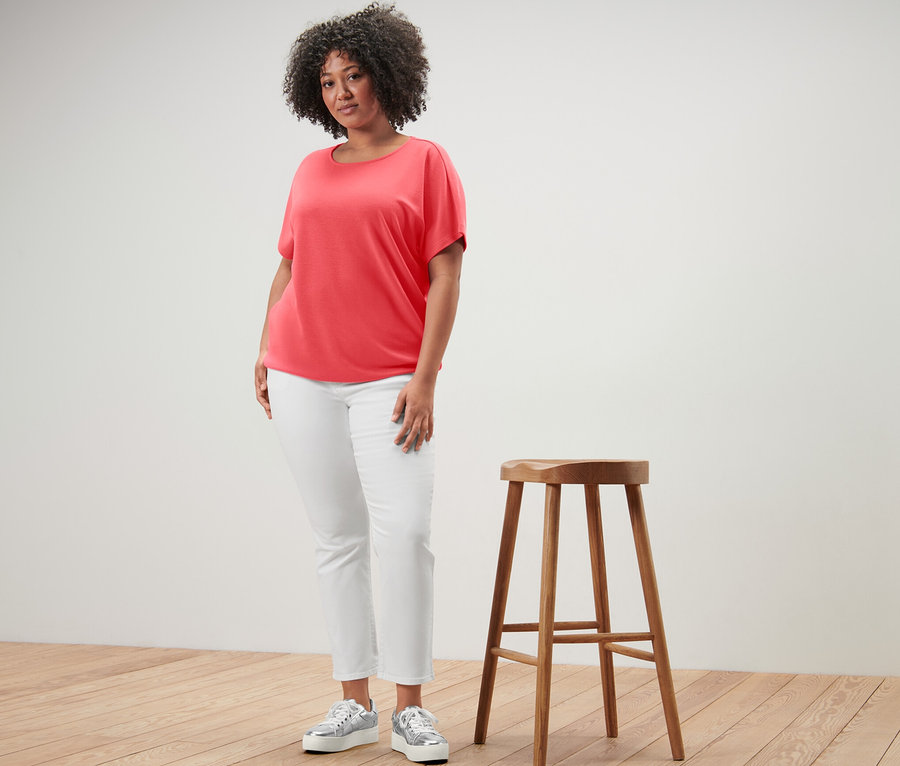 Frau steht neben einem Holzhocker und trägt ein korallenfarbenes Blusenshirt.