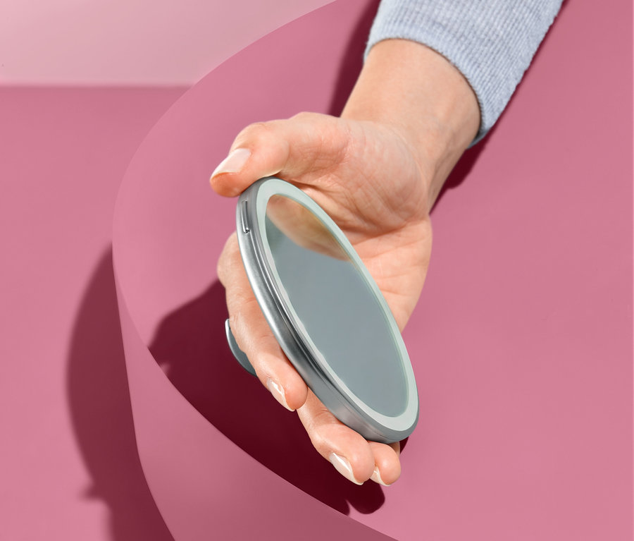 Hand hält LED-Kosmetikspiegel vor rosa Hintergrund.