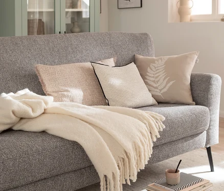 Cremefarbene SCHÖNER WOHNEN-Kollektion Wohndecke »PLENTY« über einem grauen Sofa mit drei Zierkissen, daneben ein Bücherstapel und ein hellgrüner Schrank.