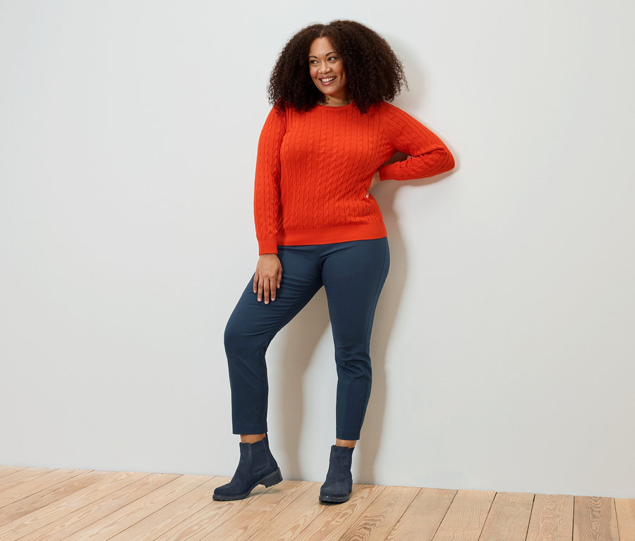 Lächelnde Frau mit lockigem Haar posiert in orangefarbenem Pullover, blauen Hosen und Stiefeln.