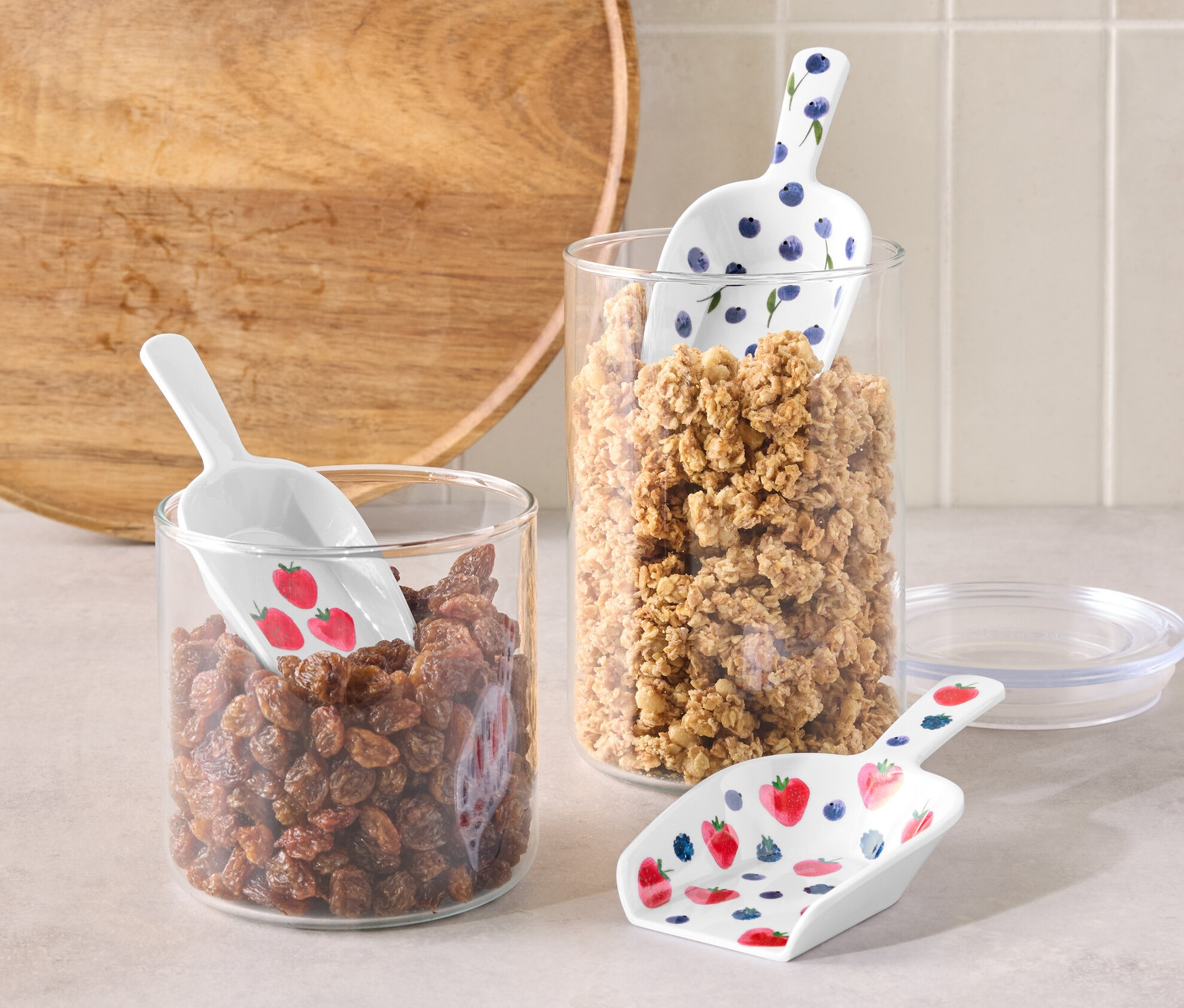 Einmachglas mit Rosinen und 3 Dosierschaufeln mit Erdbeeren und Einmachglas mit Müsli und 3 Dosierschaufeln mit Blaubeeren auf einem Tisch.