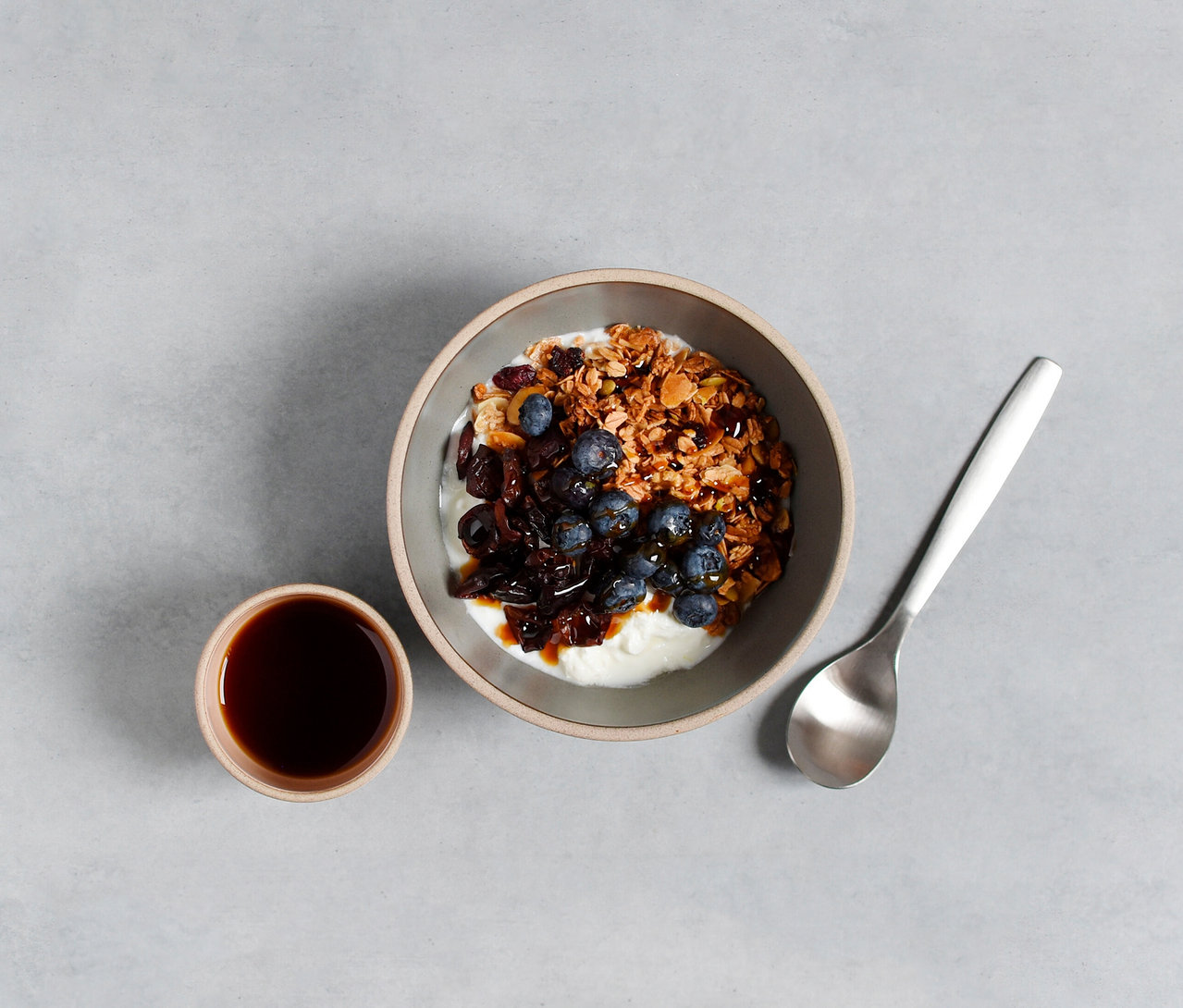Draufsicht auf eine Schale Joghurt mit Müsli, Blaubeeren und Sirup neben einer Tasse Kaffee und einem Löffel auf grauem Hintergrund.