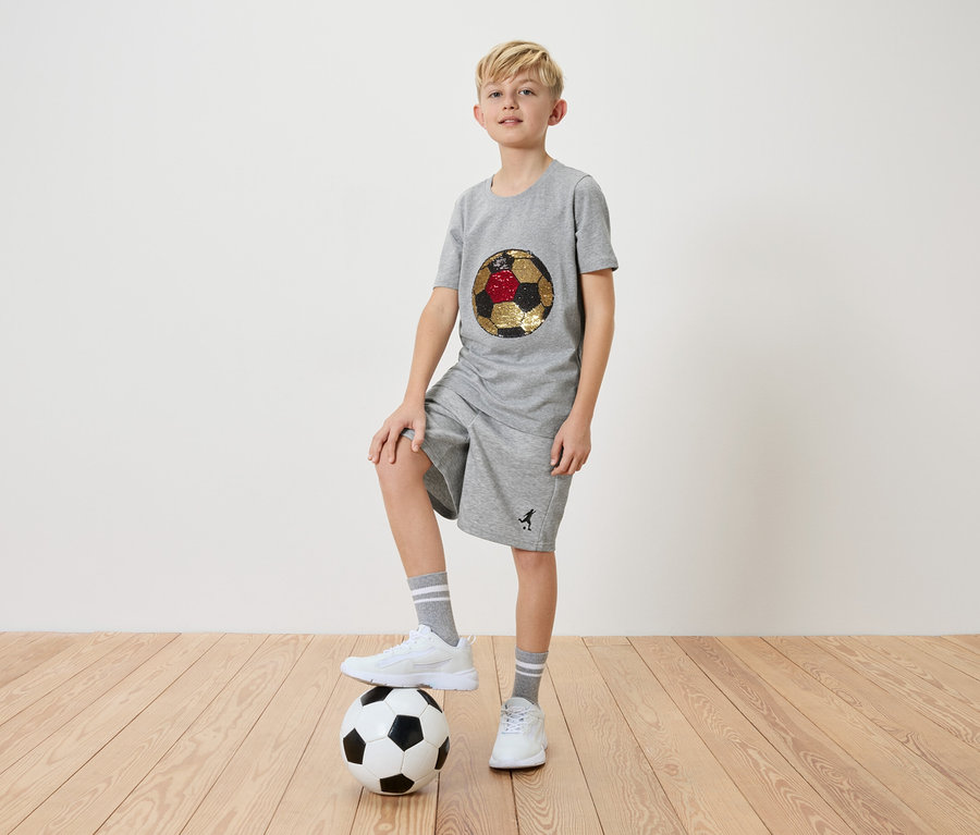 Ein blonder Junge in grauem T-Shirt und Shorts steht mit einem Fuß auf einem Fußball.