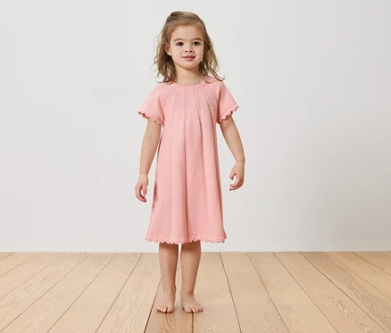 Mädchen steht barfuß auf einem Holzboden und trägt ein rosa Kleid.