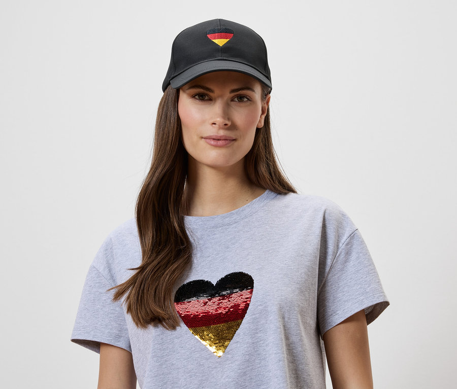 Frau mit grauem T-Shirt mit Herz in den deutschen Farben und schwarzer Kappe mit kleinem Herz in den deutschen Farben.
