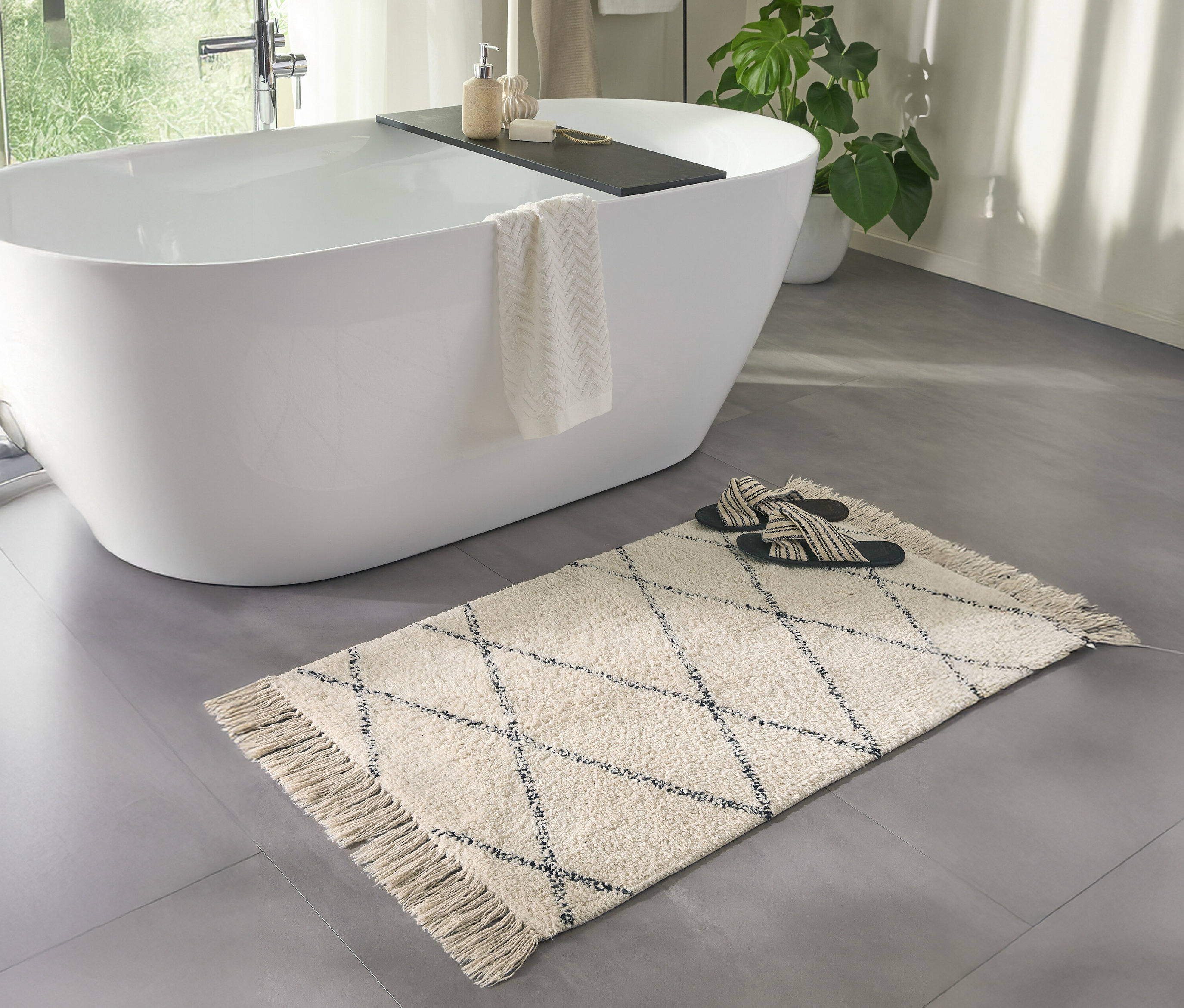 Ein Badezimmer mit einer weißen Badewanne und einer cremefarbenen benuta Badematte »Naomi« (50 x 80 cm) mit Fransen.