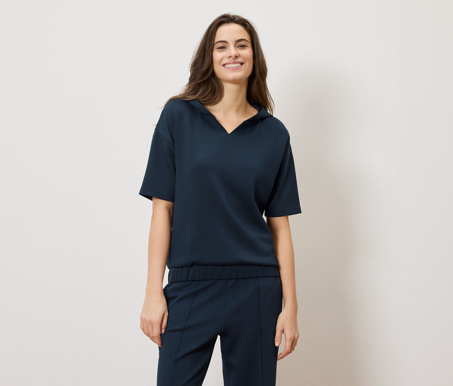 Eine Frau mit langen braunen Haaren lächelt und trägt ein dunkelblaues, kurzärmeliges Oberteil und eine Hose.