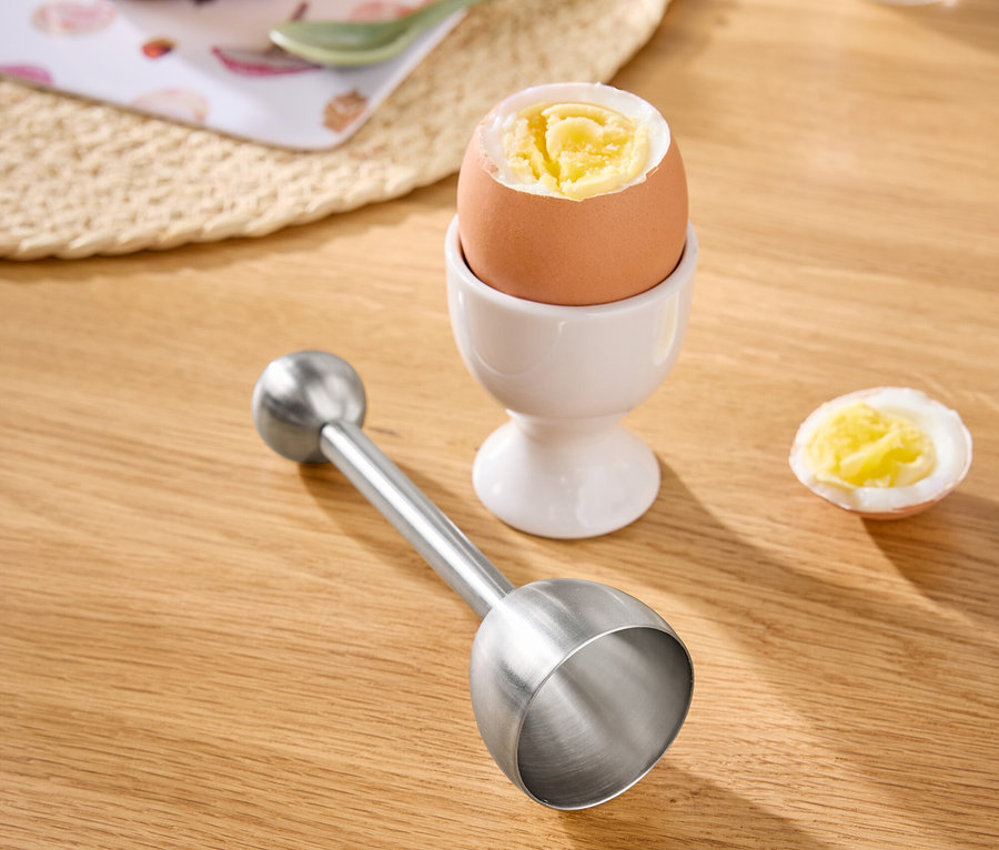 Auf einem Holztisch stehen ein Ei im Eierbecher, ein halbes Ei, ein Eierschalensollbruchstellenverursacher, ein Brot mit Käse und eine Salzmühle.