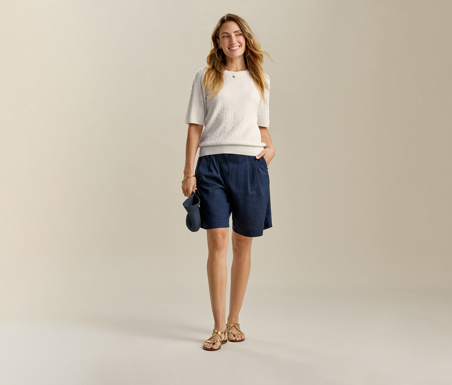 Frau in hellem Oberteil und dunkelblauen Leinen-Bermuda-Shorts, mit einer Hand in der Hosentasche.