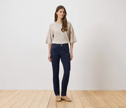 Frau mit beigem Pullover und dunkler Jeans steht auf einem Holzboden vor einer weißen Wand.