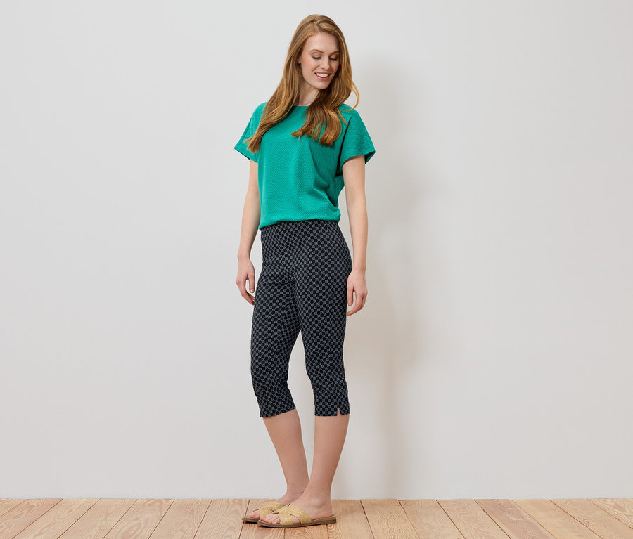 Frau mit langen, roten Haaren, die ein grünes T-Shirt und eine dunkle Caprihose mit geometrischem Muster trägt.