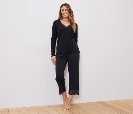 Frau in schwarzem, langärmligem Oberteil mit V-Ausschnitt und schwarzer Hose mit Spitze am Saum, vor einer weißen Wand.