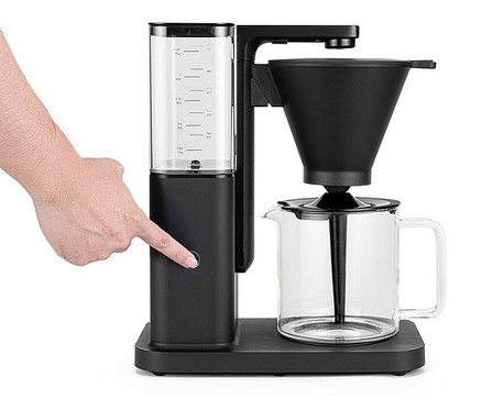 Eine Hand drückt den Knopf an einer schwarzen Kaffeemaschine mit Glaskanne und transparentem Wasserbehälter.