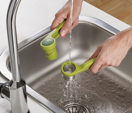 Eine Person spült mit einem grünen Küchenutensil unter einem Wasserhahn in einem Spülbecken.