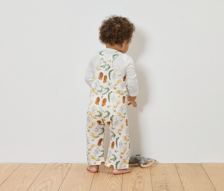 Kleinkind von hinten in Musselin-Latzhose mit Tierprint vor weisser Wand.