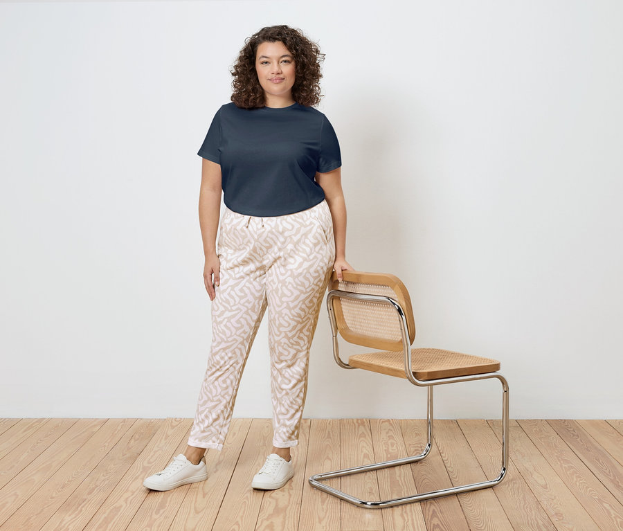 Eine Frau mit lockigem Haar steht neben einem Stuhl mit Holzrahmen und Rattanbespannung. Sie trägt ein marineblaues T-Shirt, gemusterte Hosen und weiße Turnschuhe.