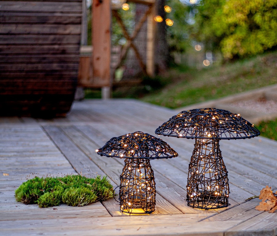 Auf einer Holzterrasse stehen zwei pilzförmige, schwarze Drahtleuchten mit warmweißen Lichtern neben Moos.