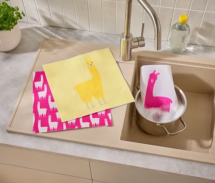 Gelbes und pinkes Tuch mit Lama-Motiv liegen neben einem Spülbecken, in dem ein Topf mit einem Tuch mit Lama-Motiv steht.