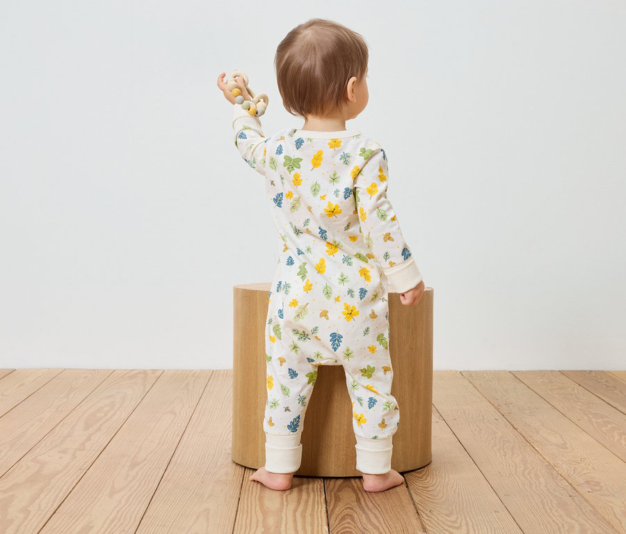 Ein Kleinkind in einem Blätter-Overall steht mit dem Rücken zu einem Holzschemel und hält ein Holzspielzeug in der Hand.