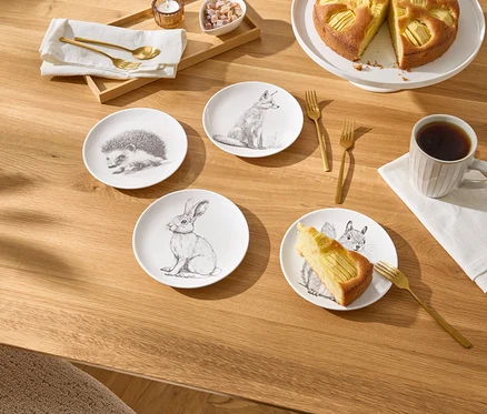 Auf einem Holztisch stehen vier kleine Teller mit Tierzeichnungen, ein Stück Apfelkuchen, eine Tasse Kaffee und goldenes Besteck.
